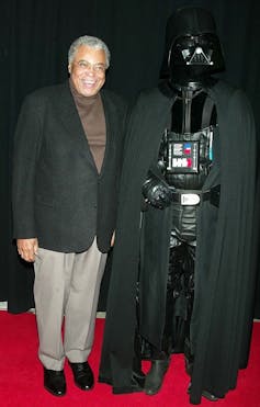 Older Black man wearing suit jacket poses on red carpet next to sinister-looking, costumed person wearing a black mask and black cape.